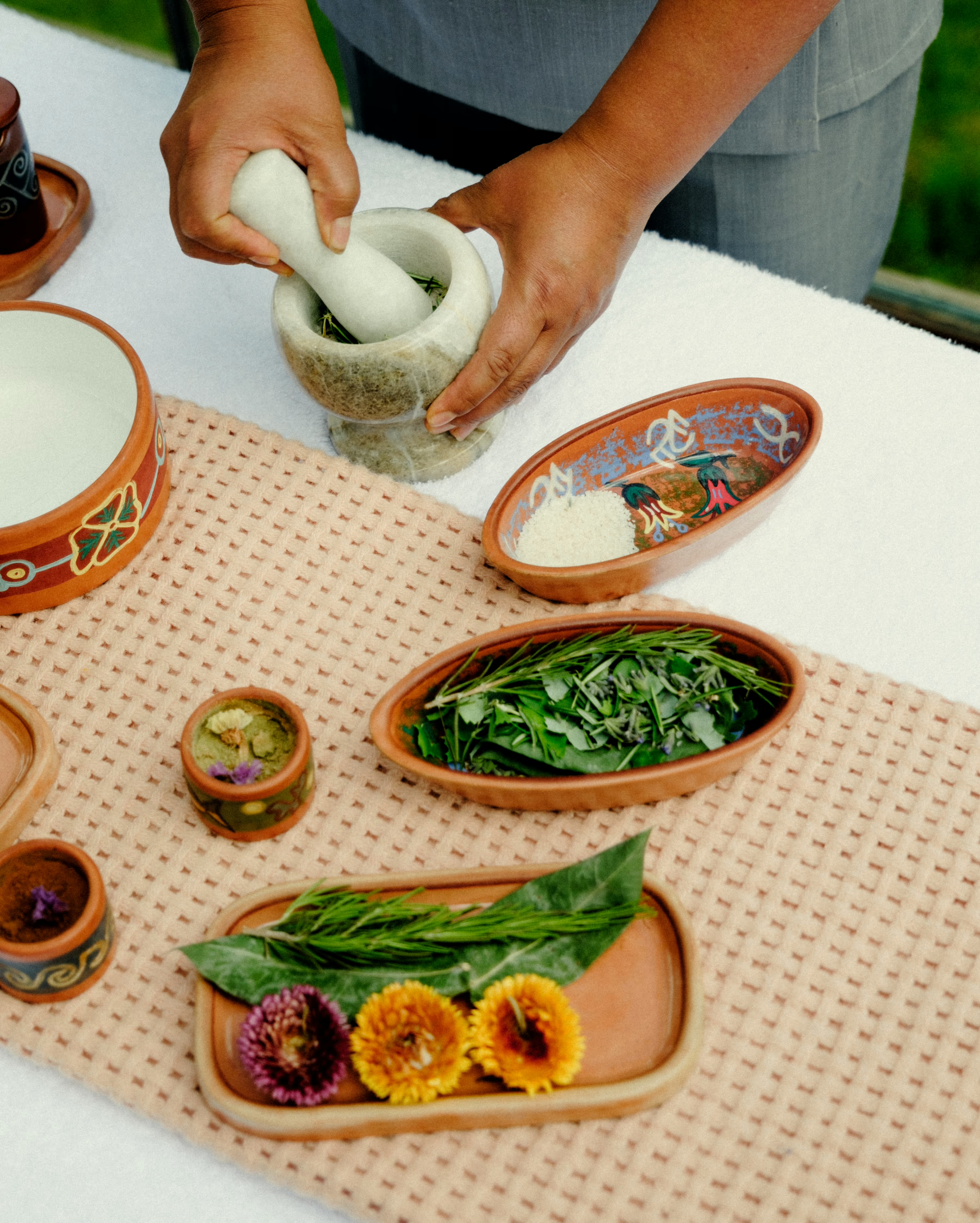Hands grinding herbs in a stone mortar beside painted dishes of Andean flowers, grains and greens